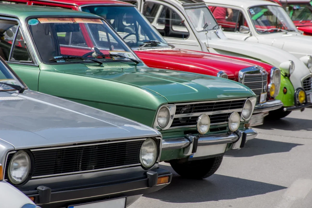 Row of classic cars parked side by side at an outdoor car show, including a silver hatchback, a green sedan, a red Mercedes-Benz sedan and white vintage compacts